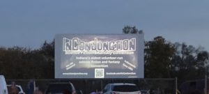 A drive-in movie screen rises up in front of a tree line at twilight. On it is a slide for InConJunction Science Fiction & Fantasy Convention - its banner-like logo featuring InConJunction in block letters, with other details (including inconjunction.org and a QR code leading to it)